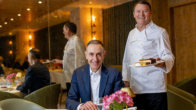 Elie Maalouf, CEO of IHG Hotels and Resorts, in a suit, sits at a restaurant table with pink flowers. Chef Theo Randall stands behind him, holding a plate of desserts.